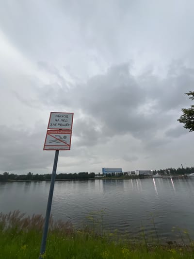 The image depicts a gloomy, overcast day at the Neva River in the city of Imeni Sverdlova, Russia. A metal pole supporting two red and white signs is visible in the foreground. The top sign reads "ВЫХОД НА ЛЁД ЗАПРЕЩЁН" (Exit onto the ice is prohibited). The bottom sign features a graphic of a person swimming with a diagonal line through it, indicating that swimming is forbidden. Below this graphic, smaller text states "КУПАНИЕ ЗАПРЕЩЕНО" (Swimming is prohibited) and "ПРИ НЕСТАЧНЫХ СЛУЧАЯХ НА ВОДЕ ЗВОНИТЕ 01, СОТОВЫЙ 112" (In case of emergencies on the water, call 01, mobile 112).
The river stretches across the mid-ground, with a distant shoreline featuring a line of trees. On the opposite bank, several industrial buildings, including one with a distinct blue and white facade, are visible. The water reflects the cloudy sky and the lights from the buildings. The foreground consists of green grass and some taller, dry vegetation. A few small yellow flowers are scattered among the grass. The overall atmosphere is somber due to the cloudy weather.