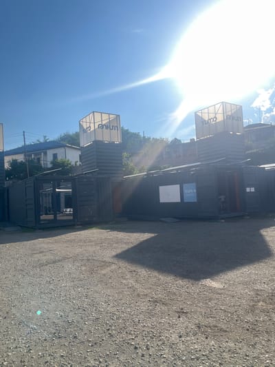 This image captures an outdoor scene in Gladzor, Armenia, on a bright, sunny day. The sun's glare is prominent, indicating it's likely late morning or early afternoon. The setting appears to be an open area with a gravel or dirt ground, possibly a parking lot or yard, adjacent to some buildings.

The main subjects are several stacked shipping containers, some of which have been repurposed as structures. One container, prominently displayed, features a white sign with a latticework frame, displaying the word "uniter" in a stylized font. Another similar sign is visible lower down. On the side of a dark grey container, there are two smaller signs; one is a blue square with white text and a logo, and the other is a white square with a line drawing. The containers are dark grey and appear to be used as modular buildings or perhaps storage units.

Through the gaps in the containers and in the background, glimpses of residential buildings and lush green trees can be seen, suggesting a semi-urban or suburban environment. The sky is a clear, vibrant blue with a few wispy clouds. The overall impression is one of a functional, possibly commercial or community space, utilizing repurposed materials. There are no people visible in the image, and the focus is on the structures and the environment. The strong sunlight casts a distinct shadow of one of the containers across the ground.