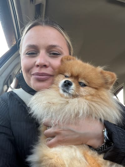 The image depicts a female individual and a Pomeranian dog inside a vehicle. The specified location is Povarenki, Russia. The woman, positioned on the left side of the frame, has light-colored hair pulled back and is wearing a dark, textured garment, secured by a light-colored seatbelt across her chest. She has light-colored lipstick and is looking directly at the camera with a slight smile. Her left arm supports the dog, and a metallic watch with a patterned band is visible on her left wrist. The dog, a small breed with fluffy, predominantly orange-brown fur and lighter cream-colored underparts, is held against the woman's chest, facing the camera. It has dark eyes and a dark nose, and its expression is neutral. The background shows parts of the car's interior, including the headliner and a window frame on the left, indicating an indoor vehicular setting. The composition suggests a close-up selfie captured within the moving or stationary vehicle.