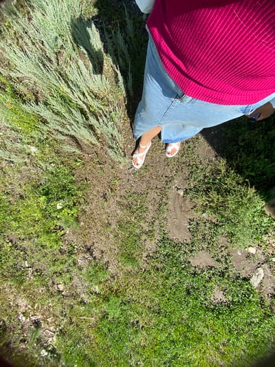 A person stands in a grassy area with sparse patches of dry dirt. They are wearing a pink ribbed sweater and a light blue denim skirt. White sandals with a strap across the toes are on their feet. The scene is outdoors, with sunlight casting shadows on the ground. The surrounding vegetation consists of green grass and tall, wispy plants. The location is the city of Yuldybayevo, Russia.
