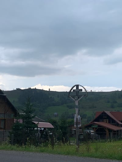 The image depicts a rural mountainous landscape in Lazeshchyna, Ukraine, under an overcast sky. In the immediate foreground, a dark road surface is visible, bordered by a strip of green grass and weeds. A chain-link fence runs horizontally across the mid-ground, behind the vegetation.

The central subject is a tall, weathered wooden crucifix, positioned to the right of the scene's vertical center. It features a carved or painted figure of Jesus Christ, adorned with a semicircular arch above the head of the corpus and an embroidered rushnyk (traditional Ukrainian towel) draped around its lower section.

To the left of the crucifix, partially obscured by the fence and a small conifer tree, is a house with a dark roof and light-colored siding. To the right of the crucifix, another house with a reddish-brown tiled roof and wooden construction, including an extended sheltered area, is visible behind the fence. A light-colored vehicle with a red cover is partially visible behind the fence, near the right house.

The background is dominated by a large, green mountain range. The lower slopes of these mountains are densely covered with dark green trees, while the upper sections feature open grassy areas with scattered individual trees. A subtle line of lighter clouds or mist is present along the distant mountain ridges. The sky occupies the upper half of the image and is completely covered by multiple layers of grey clouds, indicating an absence of direct sunlight. No people, specific actions, or interactions are visible.