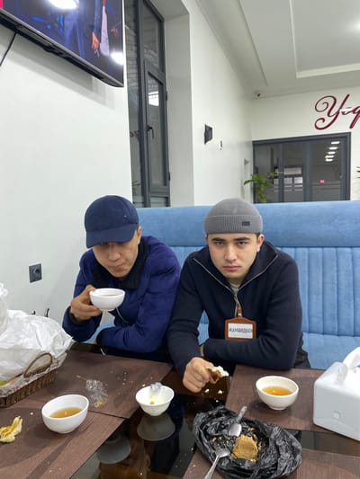 Two men sit at a glass table eating food. One man wears a grey beanie and a name tag written in a Cyrillic language, holding a piece of cake. Next to him is a black trash bag with dessert remains. The other man wears a blue baseball cap, holding a white cup. The table has three white cups, some with a yellow liquid, a plastic bag, dessert pieces, and silverware. Behind them is a blue bench. A television and windows can be seen in the background.