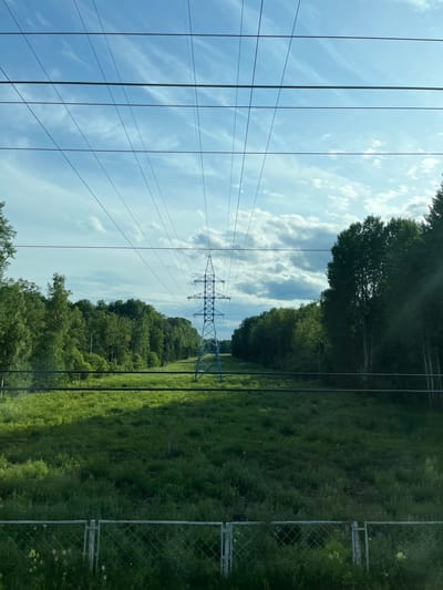 The image depicts a pastoral scene with a dominant power line pylon and its associated wires stretching across the frame. The perspective is from ground level, looking upwards and slightly to the side. The sky is a bright blue with scattered white clouds, suggesting a clear and sunny day.
To the left of the power line, several taut wires run parallel to each other, creating a strong linear element. The power line pylon, made of metal lattice, stands tall in the middle ground, supporting the overhead cables.
To the right of the pylon, a vast expanse of lush green grass and vegetation stretches into the distance, suggesting a rural or undeveloped area. A chain-link fence runs along the right edge of the frame, bordering the grassy field. Beyond the field, a dense line of green trees forms a natural backdrop. The light suggests it is daytime, possibly late morning or early afternoon.
The scene evokes a sense of open space and the presence of infrastructure within a natural landscape. The location context provided is Zhary, Russia, which aligns with the general description of a rural or semi-rural setting with power infrastructure. There is no visible text in the image.