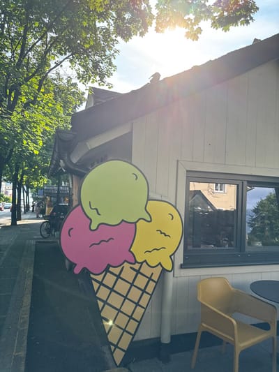 A large, colorful ice cream cone sign is mounted on the side of a building, situated in Bled, Slovenia (coordinates: 46.368, 14.111). The sign features a cone with three scoops of ice cream, depicted in green, pink, and yellow. The cone is propped against a white building with a window. A yellow chair and table are in the foreground. The scene is captured from a street-level perspective.  Sunlight shines through a tree, illuminating the sign and the side of the building. The building has a wooden roof and is decorated with large,  white siding boards. The building's window frames are dark gray. There is a sidewalk in the foreground and a tree in the background. The tree casts a shadow on the sidewalk. There are several people walking in the distance, but they are blurred and indistinct. The building is located in a residential area.  The scene is peaceful and serene, and the sign creates a sense of whimsy.