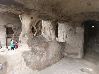 This is a photograph of an interior cave dwelling in Uplistsikhe, Georgia. The dwelling is carved into rock and features rough, uneven walls and ceiling. Several openings or doorways are visible, some arched. In the foreground, the floor is sandy and uneven, with depressions or carved out areas.
On the left side of the image, three individuals are present. A young boy in a light-colored t-shirt is standing and looking to the left. Behind him, two women are conversing. One woman wears a green shirt and dark pants, and the other wears a black tank top and a red skirt.
The environment appears to be a historical or archaeological site, likely a preserved cave complex. The lighting is natural, with light filtering in from openings, creating shadows and highlighting the texture of the rock. The overall impression is one of a subterranean, ancient living space.