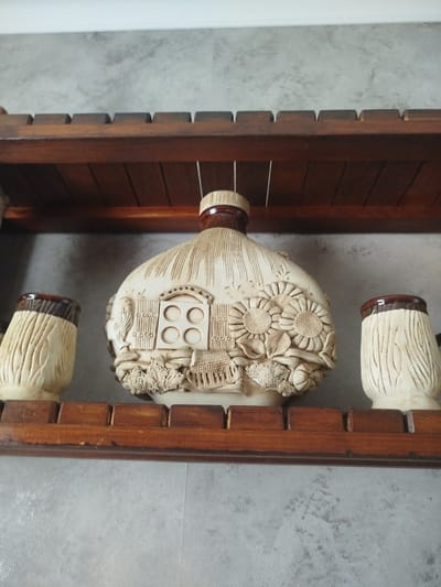 Two cream-colored, carved ceramic mugs with brown rims flank a larger, cream-colored, carved ceramic bottle on a wooden shelf. The bottle is decorated with a rustic scene featuring a fence, windows, sunflowers, and other floral elements. The shelf is made of dark wood and is mounted against a gray, textured wall. The overall impression is of a decorative display of artisanal pottery.