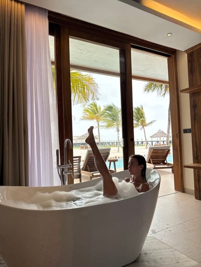 A woman relaxes in a white, oval bathtub filled with bubbles. She is facing the left of the frame, and her right leg is visible outside the tub. A large window with a wooden frame behind the tub shows a tropical outdoor scene. Palm trees, lounge chairs, and a glimpse of a pool can be seen beyond the window. White curtains hang partially over the bottom of the window.