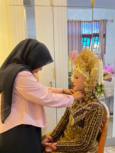 In a room in Peudaya, Indonesia, a woman wearing a black hijab and a pink long-sleeved shirt is adjusting the jewelry of another woman who is dressed in a traditional, ornate golden wedding outfit. The bride is seated, wearing a large, intricate gold headpiece adorned with white and green floral accents. Her dress is richly decorated with gold sequins and beads. The bride's hands, visible in the foreground, are decorated with henna. The scene appears to be in a dressing room or preparation area, as evidenced by the presence of mirrors and clothing racks, with a window covered by curtains visible in the background reflection.