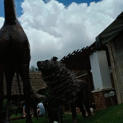 The scene depicts an outdoor area in Mweka, Tanzania, observed under a partly cloudy sky. In the foreground and midground, two prominent dark statues are visible. On the left, the torso and legs of a giraffe statue are partially cropped. Centered to its right is a dark, stylized statue of a lion, featuring a textured mane and a gaze directed upward and left. A potted plant is positioned at the base of the lion statue. In the midground, two individuals are seated on green grass, partially obscured by the statues; one wears a light shirt, the other a darker one. The background features architectural elements: a building with a thatched roof behind the statues and a structure on the right with a white pillar, a brown brick base, and an exposed wooden rafter roof section. A small, light-colored sign on a post is visible on the far right. Daylight illuminates the scene, casting shadows.