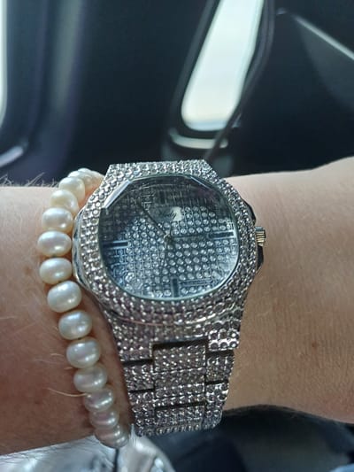 The image shows a close-up of a person's wrist adorned with a pearl bracelet and a diamond-encrusted watch. The watch has a metallic band and a faceted bezel. The dial of the watch appears to be covered in small, reflective square patterns, and a brand name, "Hedy," is visible in the center. The background is dark and out of focus, suggesting an indoor or vehicle setting. There is no indication of the location within the city of Maiaky, Ukraine.
