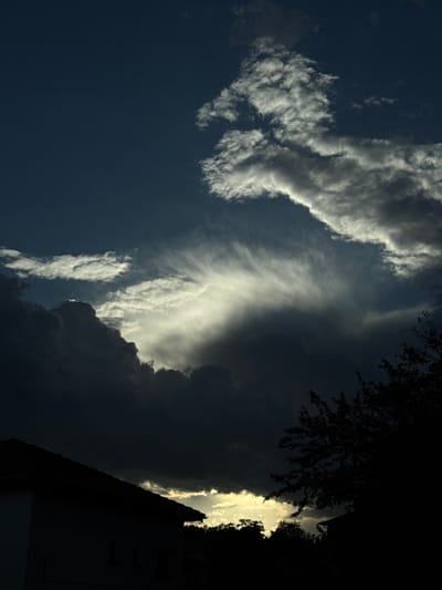 The image shows a sky scene in Zhaltusha, Bulgaria. Cumulus clouds dominate the view, contrasted against patches of dark blue sky. A building and the silhouette of a tree occupy the right side of the frame. Sunlight is seen streaming behind the clouds, creating a dramatic lighting effect. The image is oriented vertically, with the building positioned at the bottom right corner.