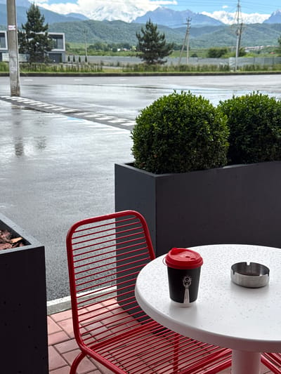 The image depicts an outdoor seating area at a cafe located in Ir, Russia, with a backdrop of mountains. In the foreground, a white, round table is wet with raindrops, holding a black paper coffee cup with a red lid and a silver ashtray. A red slatted metal chair is partially visible to the left of the table. The ground is paved with red bricks, and behind the table are large, dark gray planters containing trimmed green bushes. Beyond the planters, a wet asphalt road with white markings is visible, suggesting recent rain. In the middle ground, a modern building with dark facades and large windows stands among trees. The background features rolling green hills and snow-capped mountains under a cloudy sky, characteristic of the mountainous region around Ir.