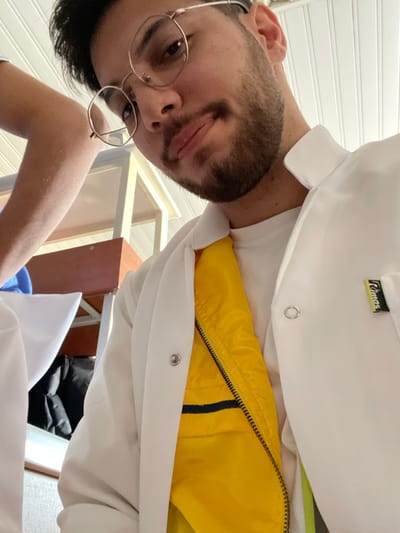 A close-up, low-angle image depicts a young adult male in قرية الضبية, Syria, engaged in a self-portrait. He has a light skin tone, dark hair, and a short beard and mustache. He is wearing round, gold-rimmed eyeglasses. The male is dressed in a white professional coat or lab coat with the lapels open, revealing a bright yellow garment or lining underneath, which is partially zipped. A white round-neck undershirt is visible beneath the yellow layer. A black and yellow tag with the word "Rimas" is affixed to the right chest area of the white coat. Metal snap fasteners are visible on the coat's front opening. His expression is neutral to slightly smiling. To the left, a portion of another individual's arm, also in a white sleeve, is visible. The background indicates an indoor setting, likely a classroom or laboratory, with light-colored shelving or furniture, and a white paneled ceiling. A dark object, possibly a bag, is partially visible beneath the furniture in the background.