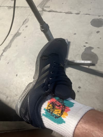 A close-up, overhead shot shows a person's left foot wearing a dark blue Adidas sneaker and a white sock with a colorful graphic design of a skull with a crown. The sneaker is laced and appears to be made of mesh material. The sock has abstract splotches of turquoise, pink, and yellow around a black skull graphic. The foot is resting on a light-colored, cracked concrete surface, possibly a sidewalk or pavement, with several dark, wet patches. A metal pole and a metal bar are visible in the background, casting shadows on the concrete. The scene appears to be outdoors in an urban environment, specifically Bogucin, Poland.
