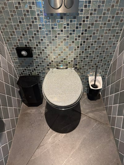 The image shows a top-down view of a modern toilet in a bathroom, possibly located in Amstelveen, Netherlands. The toilet is white with a speckled, shimmering seat and lid. To the left of the toilet is a black waste bin. To the right is a white toilet brush holder with a black toilet brush. The walls are covered in a mosaic of small, iridescent square tiles in shades of blue, green, and gray. The floor appears to be made of large, dark gray tiles with a subtle veining pattern. A metal flush plate is visible at the top of the image, mounted on the tiled wall above the toilet. The lighting creates a shadow of the toilet on the floor. There are no people or animals in the image.