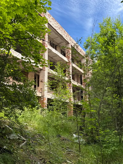An abandoned, multi-story brick building stands partially obscured by dense green foliage. The building's structure is exposed, with concrete floors and exposed brick walls. Some windows are present, but most are empty openings. The surrounding environment is overgrown with tall grass and trees, suggesting a neglected and possibly remote location within the city of Interkompleks, Russia. The sky above is blue with scattered white clouds. The ground in the foreground is littered with debris, including what appears to be fallen wooden beams and other construction materials, all covered in vegetation.