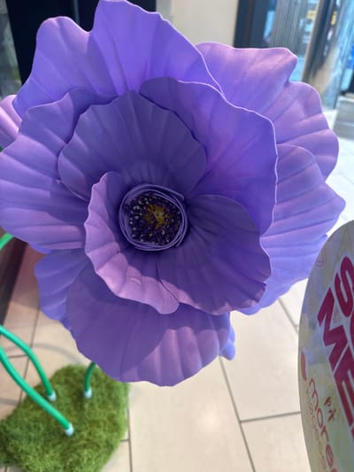 A large, artificial purple poppy is the central focus of this close-up, indoor shot. The flower's petals are layered and have a textured appearance, with prominent veins running through them. The center of the flower features a dark, circular area with yellow stamen-like protrusions and small, purple dots. The flower is supported by green wire stems that emerge from a small, green, moss-like base. In the background, a circular object with red and white text is partially visible on the right side of the image. The flooring appears to be light-colored tile. The overall impression is of a decorative item, possibly for a shop or event. The image is taken in Slitu, Norway, a town known for its historic railway station and proximity to nature.