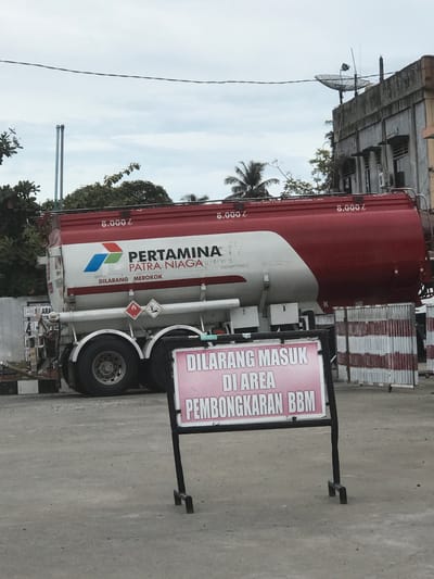 The image shows a Pertamina Patra Niaga fuel tanker truck parked on a concrete surface. A sign is standing beside it that reads "DILARANG MASUK DI AREA PEMBONGKARAN BBM" (meaning "No entry to the fuel unloading area" in Indonesian). The tanker is red and white with the Pertamina logo and the phrase "DILARANG MEROKOK" ("No Smoking"). The text "8.000 l" is written on the tanker three times. Behind the truck, there is a building with a satellite dish attached. There are trees and other vegetation visible as well. The sky is overcast.