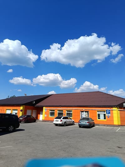 In Shishkin Les, Russia, a bright orange and yellow building with a brown metal roof stands under a clear blue sky dotted with fluffy white clouds. The building appears to be a commercial establishment, possibly a store or office, with multiple windows and doors visible. A parking lot in front of the building is occupied by several cars, including a dark SUV, a maroon sedan, a white sedan, and a grey sedan. A parking sign with a blue "P" and a wheelchair symbol is mounted on the building's exterior. The overall scene depicts a typical day in a suburban or commercial area.