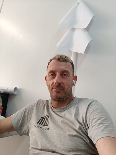 A candid medium shot shows a fair-skinned adult male with light brown hair, styled in a short, spiky cut with blond highlights. He is looking directly at the camera with blue eyes and a slight smile. He has stubble on his chin and a light beard. He is wearing a light gray t-shirt with a black logo on the left chest that appears to be an abstract design of vertical lines. He has a white wireless earbud in his right ear. He is seated in front of a white wall, which is illuminated from the left, casting subtle shadows. Two pieces of white paper are folded and attached to the wall behind him. To his left, partially visible, is a dark surface with a rolled-up document and the top of a red bottle. The image appears to have been taken indoors, in an office or workshop setting, possibly in Katowice, Poland, given the implied context of the image request.