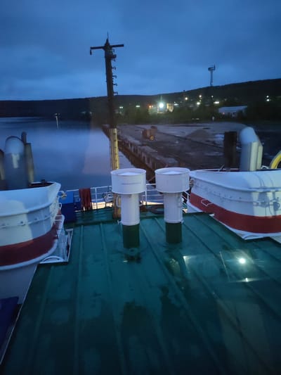 The image captures a twilight or early night scene from the deck of a vessel moored at a dock in Medvezhyegorsk, Russia. The deck, appearing wet and reflecting light, features two white and red lifeboats or small tenders, and two tall, white cylindrical ventilation structures. One of these structures prominently displays the Cyrillic text "АККУМУЛЯТОРНАЯ," which translates to "BATTERY ROOM" or "ACCUMULATOR." A mast-like structure rises from the vessel on the left.

Beyond the vessel, a calm body of water, possibly a lake or bay, stretches towards a wooden pier. The water's surface is dark and reflective, with a light mist or fog hovering above it. Along the distant shore, a settlement is visible, nestled against a dark, forested hill. Numerous artificial lights from buildings and streetlights dot the landscape, creating warm glows against the cool blue, overcast sky. A tall, industrial-looking tower stands on the hillside, overseeing the settlement.

The time of day is clearly evening or early night, characterized by the deep blue sky and reliance on artificial illumination, suggesting either dusk, early morning, or the prolonged twilight of northern latitudes. No people are visible, contributing to the quiet and serene atmosphere. The overall impression is one of peaceful stillness at a northern harbor.