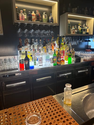 The image shows a well-stocked bar, likely in Giugliano in Campania, Italy, given the context provided. The bar features multiple shelves displaying a variety of liquor bottles, including brands like Opihr, Gin, Baileys, and Havana Club. Wine glasses are hung upside down from a rack above the counter, suggesting a prepared setup for serving drinks. In front of the shelves, a row of stacked glasses is visible, along with various smaller bottles and mixers. The counter itself is made of stainless steel, and a small digital display reading "5" and "06" is present on one of the cabinets below. Further to the right, a small, empty glass bottle with a white cap sits on the counter, next to a metal sink. The overall environment appears clean and organized, typical of a commercial establishment focused on beverages. There are no people visible in the image.