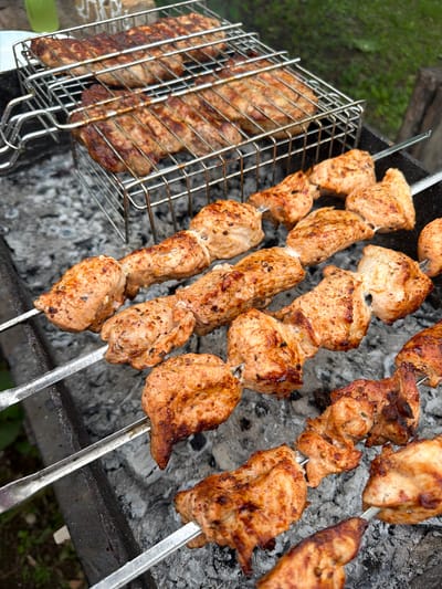 This is an overhead shot of a barbecue grill with several skewers of seasoned meat cooking over hot coals. The meat appears to be chicken or pork, cut into bite-sized cubes and threaded onto metal skewers. Some of the meat is browned and slightly charred, indicating it is nearing completion. To the left of the skewers is a metal grilling basket containing more pieces of meat, possibly sausages or ribs, also showing signs of grilling. The grill is set on a rough surface, likely outdoors, with hints of greenery in the background suggesting a garden or yard. The scene evokes a feeling of a casual outdoor meal, possibly a summer gathering. There is no visible text in the image.