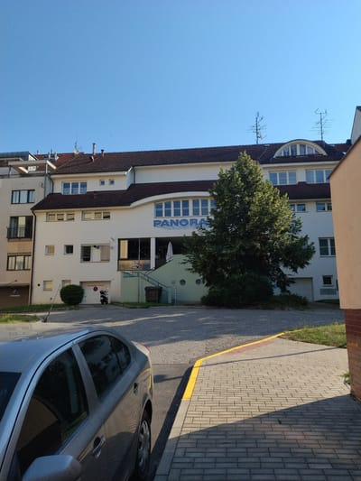 This image depicts a clear, sunny day in Hluboká nad Vltavou, Czechia. In the foreground, a silver sedan is parked on a paved driveway. The car is angled towards the right of the frame, with its driver's side door and windows clearly visible. Beyond the car, a building with a white facade dominates the scene. The building has multiple floors and a brown tiled roof. A large green tree stands in front of the building, partially obscuring a section of its facade. Above the entrance to the building, the word "PANORAMA" is displayed in large blue letters. A set of stairs leads up to the entrance, which appears to be a business with a window displaying the word "Bistro." To the left of the building, a smaller, multi-story structure with a beige facade can be seen. The background is a clear blue sky. The environment is urban, with paved surfaces and some greenery. There are no people visible in the image.