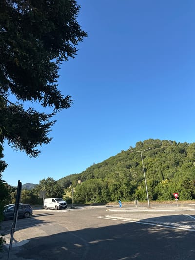 The image depicts a rural intersection in Générargues, France, under a clear blue sky. A dense, green hillside covered in trees dominates the background. In the foreground, a paved road curves to the right, forming what appears to be a roundabout. A white van is parked on the side of the road, and a dark grey car is parked to its left, partially obscured by foliage and a signpost. A modern streetlight stands in the center of the road. Traffic signs are visible: a blue circular sign with a white arrow pointing right indicates traffic flow, and a red and white triangular sign with a red border signifies a yield. The overall scene suggests a quiet, sunny day in a village setting.