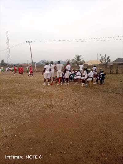 A group of people wearing white uniforms and green caps are sitting and standing on a grassy field. In the background, another group of people in red and green uniforms are playing a game. A transmission tower and power lines are visible in the distance, along with a small building and some trees. A brick wall surrounds the field. The ground is mostly dirt and dry grass.