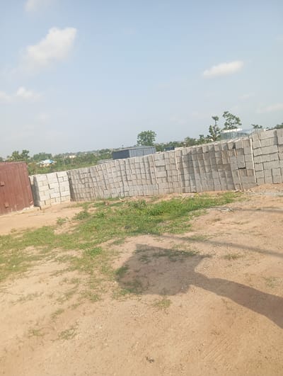 The image depicts an outdoor scene in Tasha, Nigeria, likely a construction or material storage area. A large stack of concrete blocks is prominently featured, arranged in neat rows. To the left of the blocks, a tall, reddish-brown metal structure, possibly a shipping container or storage unit, stands. The ground is covered in a mixture of sand and sparse green grass. In the background, trees and some buildings are visible, suggesting a developed area. The sky is partly cloudy with a light blue hue. A long shadow stretches across the ground in the foreground, indicating the presence of an unseen object or structure casting it.