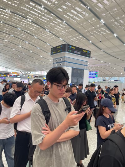 This is a photograph taken in a brightly lit, modern indoor space that appears to be an airport or train station in China. The ceiling is a prominent feature, with a complex, arched design of white panels and integrated lighting, creating a grid-like pattern that stretches into the distance.
The scene is populated by a crowd of people, many of whom are standing or sitting, looking at their phones or waiting. The people are diverse in age and attire, suggesting a busy travel hub. In the foreground, a young man with glasses is looking towards the camera, holding a smartphone in his hand. To his left, an older man in glasses is also looking down. Further back, more individuals and groups are visible, some with luggage.
Several signs are visible in the background, some displaying what appear to be departure or arrival information in a combination of Chinese characters and numbers, such as "10-20e" and "1E-9e". The overall atmosphere seems to be one of waiting and transit, with no strong indication of emotion, but a general sense of busyness. The lighting suggests it is daytime, and the indoor setting implies the weather outside is not a factor. The architectural style and signage point to a contemporary transportation facility in China, likely within Longhua District.