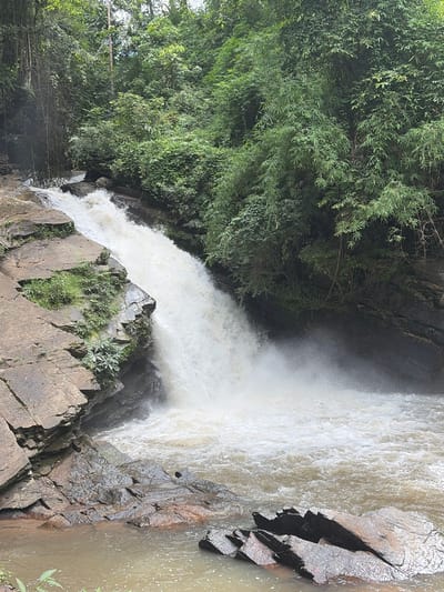 The image depicts a natural waterfall cascading down a rocky incline within a lush, verdant forest. The water is a light brown color and churns vigorously as it plunges into a pool below. The surrounding vegetation is dense, with a variety of trees and vines creating a canopy overhead. Rocks of various sizes, some covered in moss and small plants, line the sides of the waterfall and the pool. The scene appears to be a secluded natural area, typical of the undeveloped landscapes found near Ban Huai Takhrai, Thailand, known for its natural beauty and outdoor recreational opportunities. There are no people or man-made structures visible in the image.