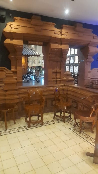 The image captures an interior view of "Kedai Tempo Doeloe" (Old Times Cafe) in Glantengan, Indonesia. The foreground features a light-colored tiled floor with a black and white decorative border. Arranged on the floor are four traditional wooden chairs, including two armchairs and two smaller stools or chairs. A large, intricately carved wooden partition, resembling a Javanese architectural gate, spans the midground, featuring multiple openings. Above the central opening, a sign reads "KEDAI TEMPO DOELOE." Through the openings, a dark-colored motorcycle, appearing to be a cruiser, is parked on a light-colored floor in an adjacent area. Beyond the motorcycle, another building structure with a traditional roofline and framed images or exhibits on a wall are visible. The background walls of the immediate space are dark, possibly blue or black, and the area is illuminated by overhead lights. No individuals are present in the scene.