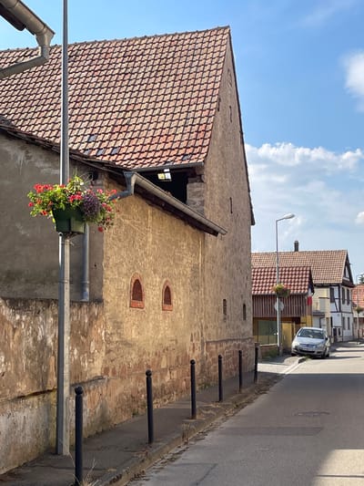The image shows a street scene in Gerstheim, France, likely taken during the daytime on a clear, sunny day with some clouds. The focus is on the side of an old stone building with a red-tiled roof. The building has two small, arched windows near the top of its wall and a hanging planter filled with colorful flowers, including red and purple blooms, attached to a pipe.
In the background, there are more buildings with red-tiled roofs, some of which appear to be houses or shops. A street runs along the right side of the image, bordered by a low wall and several black metal posts. A silver car is parked on the left side of the street. A street lamp is visible on the left side of the image, extending over the street. The overall impression is a quiet, residential or semi-commercial street in a European town.