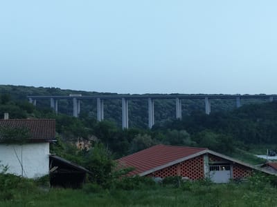 The image depicts a rural landscape near Pisanets, Bulgaria, at dusk or dawn. In the mid-ground, a prominent, long concrete viaduct with multiple tall piers spans a deep valley covered in dense, dark green deciduous forest. A light-colored truck is visible traveling on the viaduct deck, moving from right to left.

In the foreground, a grassy, overgrown field slopes gently downwards. To the left, a portion of a white, single-story house with a red tiled gable roof is visible, along with an attached wooden shed. To the right, another single-story building features a red tiled gable roof and a distinct facade constructed from reddish-brown brick with a patterned openwork design. This building has a light-colored garage door, inside which a light source is visible. Further to the right and slightly in the background, a dark vehicle is faintly visible on a distant road. The sky above is a pale, clear blue, indicating low light conditions. No individuals are discernible in the scene.