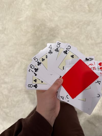 A hand holds a fan of playing cards, mostly of the suit of clubs, ranging from two to eight, along with an Ace of Clubs, five of hearts, King of hearts, and a blank red card. The background is a fuzzy, light-colored surface. A sleeve of a brown shirt is visible on the right edge of the image. The location is not discernible.