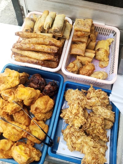 An overhead shot displays various Indonesian fritters arranged in trays and baskets. Crispy spring rolls and Martabak Telor fill the top containers. A blue tray on the left overflows with fried tofu and dark-colored tempeh. The right tray, also blue, contains fried vegetables, possibly Bakwan. Tongs rest among the fried tofu.
