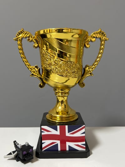 The scene, located in the city of Breznitsa, Bulgaria, displays three primary objects arranged on a flat, white surface against a uniform grey background. A prominent golden trophy, approximately 20-25 cm in height, occupies the central position. This trophy features a cup-shaped body with two ornate, curved handles, and its surface is decorated with horizontal textured bands and a central relief pattern of roses. It stands on a black, rectangular plinth. Attached to the front face of this plinth is a rectangular emblem depicting the United Kingdom's Union Jack flag. To the immediate left of the trophy's base, a small, blocky black figurine, identifiable as the Ender Dragon from the video game Minecraft, rests on the white surface, characterized by its purple eyes, gray horns, and white wing details. No individuals or active interactions are present in the static indoor environment.