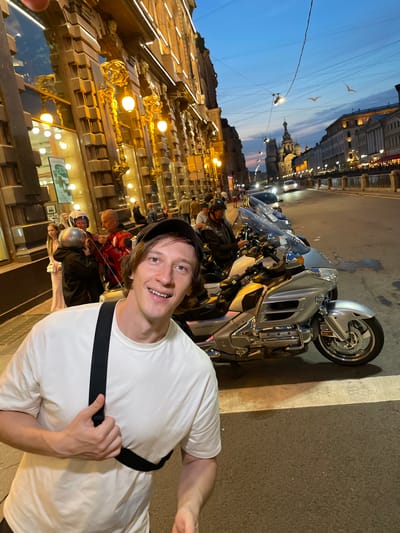 A man wearing a white t-shirt and a black baseball cap smiles at the camera while standing on a city street at dusk. Behind him, several motorcycles are parked along the sidewalk. The buildings in the background are lit, and the sky is a deep blue with hints of pink. In the distance, a church with a dome is visible. The overall scene suggests a lively urban environment with a touch of historic architecture. The city of Kokkorevo, Russia, is mentioned as the location.