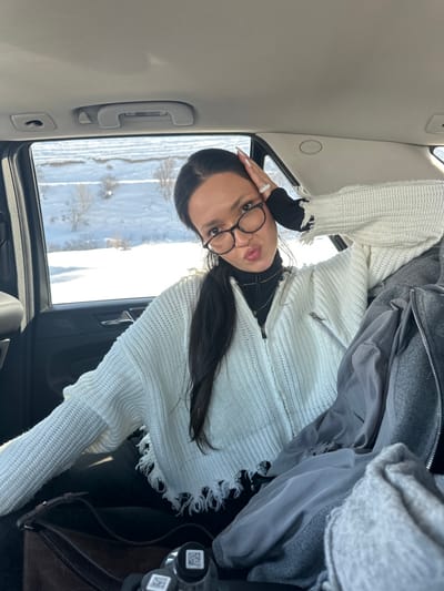 The scene is set inside a vehicle, specifically the rear passenger compartment, with a snowy exterior landscape visible through the window. The location is Jraber, Armenia.

A single female individual is present, positioned in the right rear passenger seat. She has dark hair, which is styled down, and wears round, dark-rimmed eyeglasses. Her attire consists of a black high-neck top layered under a white, chunky knit zippered cardigan featuring distressed, frayed edges at the hem and cuffs. Visible accessories include multiple rings on her right hand and long, light-colored fingernails.

The individual is actively posing for the camera, her head slightly tilted, with her right hand resting near her temple. Her lips are puckered in a distinct expression, and she maintains direct eye contact with the camera.

Within the vehicle's interior, a light-colored headliner and a dark door panel are visible. A dark-colored bag is partially observed on the seat beside her, accompanied by a pile of gray clothing. In the lower left foreground, two small, clear bottles with black lids and white QR code stickers are discernible.

Outside the vehicle, a snow-covered landscape unfolds, characterized by rolling hills or terraced terrain under a bright sky. Sparse trees or bushes are scattered across the snow-laden ground.
