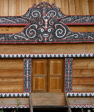 The image shows the ornate wooden facade of a traditional house in Ambarita, Indonesia. The structure features intricate carvings and decorative patterns in black, white, and red against the natural wood grain. A prominent, stylized face-like motif is central to the upper decorative element. Below this, a set of double wooden doors are flanked by vertical carved panels. Decorative borders with geometric and swirling patterns run horizontally above and below the doors and along the lower edge of the visible structure. Wooden steps lead up to the doors. The overall impression is of rich cultural heritage and detailed craftsmanship.
