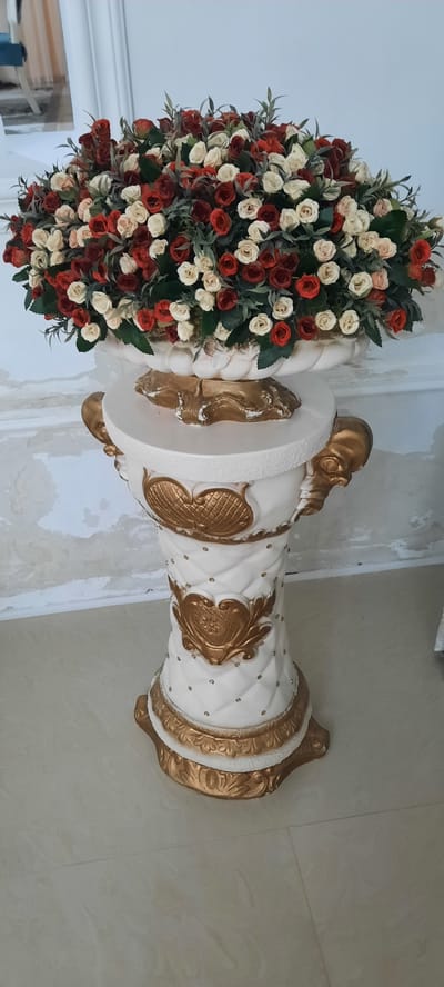 The image displays a decorative arrangement consisting of a large bouquet of artificial flowers and a ornate white and gold pedestal. The bouquet, positioned on the left side of the frame, is densely packed with red, cream, and blush-colored roses interspersed with green foliage. The pedestal, on the right, is cylindrical with a flared base and top. Its surface is textured and features raised gold decorative elements, including swirling patterns and ornate crests. Small, clear embellishments are scattered across the white surface of the pedestal. The background appears to be an indoor setting, with a tiled floor and a wall that shows signs of wear or unfinished plaster. The lighting suggests an indoor environment with ambient light, and there are no clear indicators of time of day or weather. The image is oriented vertically, as if the camera was held upright. The overall impression is of a decorative display, possibly for an event or as a permanent fixture in a room. There is no visible text in the image. The context of Gudermes, Russia is provided but not visually evident within the image itself.
