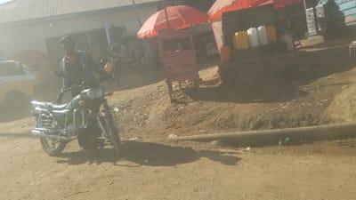 The scene is a dirt road in Kayna, Democratic Republic of the Congo, with a man standing beside a green motorcycle. He is wearing a black jacket and a dark colored hat. The motorcycle is parked on a dirt road in front of a small, red, open-air shop with a sign that reads "MINJUK SHOPS". There is a metal pipe running diagonally across the road in front of the motorcycle. In the background, there are two red umbrellas, one in the front and another in the back, with a yellow car parked partially visible behind the motorcycle. The man is looking at the motorcycle and appears to be about to leave. There are no other people visible. The sun is shining brightly. The location is near the intersection of Avenue de la Paix and Avenue des Nations Unies in Kayna.