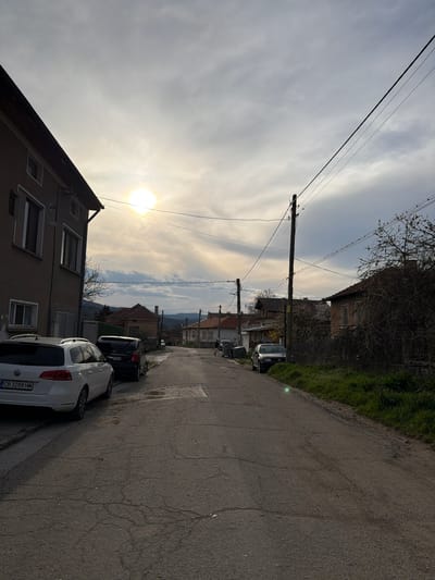 The image depicts a street scene in Popintsi, Bulgaria. The view is along a narrow asphalt road, showing cars parked on the right side. Buildings line the street, one of which is a two-story structure with visible windows. Overhead, power lines traverse the sky. The sky is overcast with visible sun. In the distance, the silhouette of hills can be discerned.