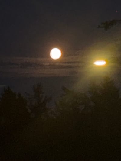 This is a nighttime photograph of the sky over Neumünster, Germany. The image features a blurry, bright circular object in the upper left quadrant, likely the moon, partially obscured by clouds. To the right of the moon, another bright, elongated, and blurry light source is visible. In the foreground, the silhouettes of trees are dark and indistinct against the night sky. The overall scene is dimly lit and appears to have been captured with a low-light setting, resulting in a lack of sharp detail.