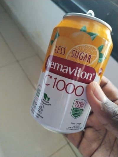 A person's hand holds an orange and white can of Hemaviton C1000 less sugar orange flavored health drink. The can has a pull tab on top and features images of oranges and leaves. Text on the can includes "Hemaviton C1000", "Less Sugar", "Total Care", "Vitamin C", "Vitamin D3", "Zinc", "Stevia Pemanis Alami", and "HALAL INDONESIA". The background appears to be an indoor setting with tiled flooring. The image location is not specified to be in the city of Lahat, Indonesia.