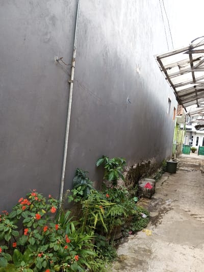 The scene depicts a narrow outdoor passage in Viengthong, Laos, situated between two structures under an overcast sky. To the left, a tall, grey, unpainted concrete wall extends from the foreground into the background, exhibiting areas of water staining and organic growth along its lower section. A silver-colored metal conduit pipe runs vertically up this wall, secured by two metal clips. Two thin wires, one blue, are attached to the wall's surface. At the base of the wall, a planter bed contains various green plants, some bearing small, orange-red flowers. The ground consists of an uneven concrete path, displaying patches of moisture and discolored areas, with a yellow piece of debris visible. Along the right side of the path, a dark green cylindrical bucket and a white open-top container are present. Above the right side of the path, a makeshift awning constructed from translucent corrugated plastic panels and a supporting frame extends over a portion of the walkway. In the background, additional buildings with windows and green gate elements are visible. No individuals are present, and no active interactions are depicted.