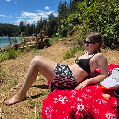 A young woman is lying on a red blanket with a snowflake pattern on a sandy bank next to a body of water. She is wearing a black bikini top and black and white patterned athletic shorts. Water droplets are visible on her skin, suggesting she has recently been in the water. She is wearing sunglasses and looking down. In the background, there are trees, a body of water, and a blue sky with white clouds. A piece of driftwood is visible on the left side of the image. A small orange plastic container is on the blanket. The image appears to be taken on a sunny day, likely in the afternoon, given the strong shadows. The presence of trees and a lake suggests an outdoor, natural setting. There is visible text on the shorts that reads "adidas".