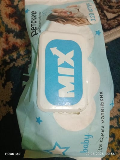The image shows a close-up of a package of baby wet wipes. The packaging is primarily light blue with white and turquoise accents. Prominently displayed on the front is the word "MIX" in large turquoise letters within a white oval shape. Above this, there is text in Russian that translates to "Children's wet wipes" and "120 pieces." A faint image of a sleeping baby is also visible on the packaging. On the right side of the package, vertically oriented Russian text reads "For the smallest ones." In the bottom right corner, a timestamp "29.06.2026 00:56" is present, along with "POCO M5" in the bottom left corner, indicating the device used to capture the image. The background consists of a patterned carpet. There are no people visible in the image. There is no indication of the city of Hisor, Tajikistan.